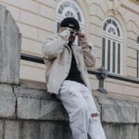 A man in fashionable casual wear, sitting outdoors against a historic building.